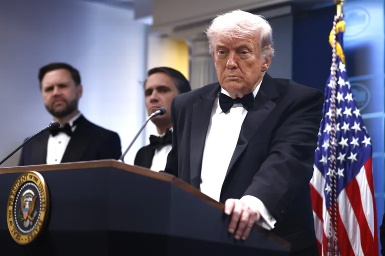 U.S. President Donald J. Trump speaks at a podium in the White House press briefing room, wearing a black tuxedo, with two administration officials standing behind him and an American flag positioned to his right.