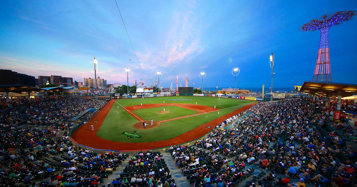 A packed Brooklyn Cyclones ballpark at sunset with fans, bright stadium lights, and amusement park rides visible beyond the outfield.