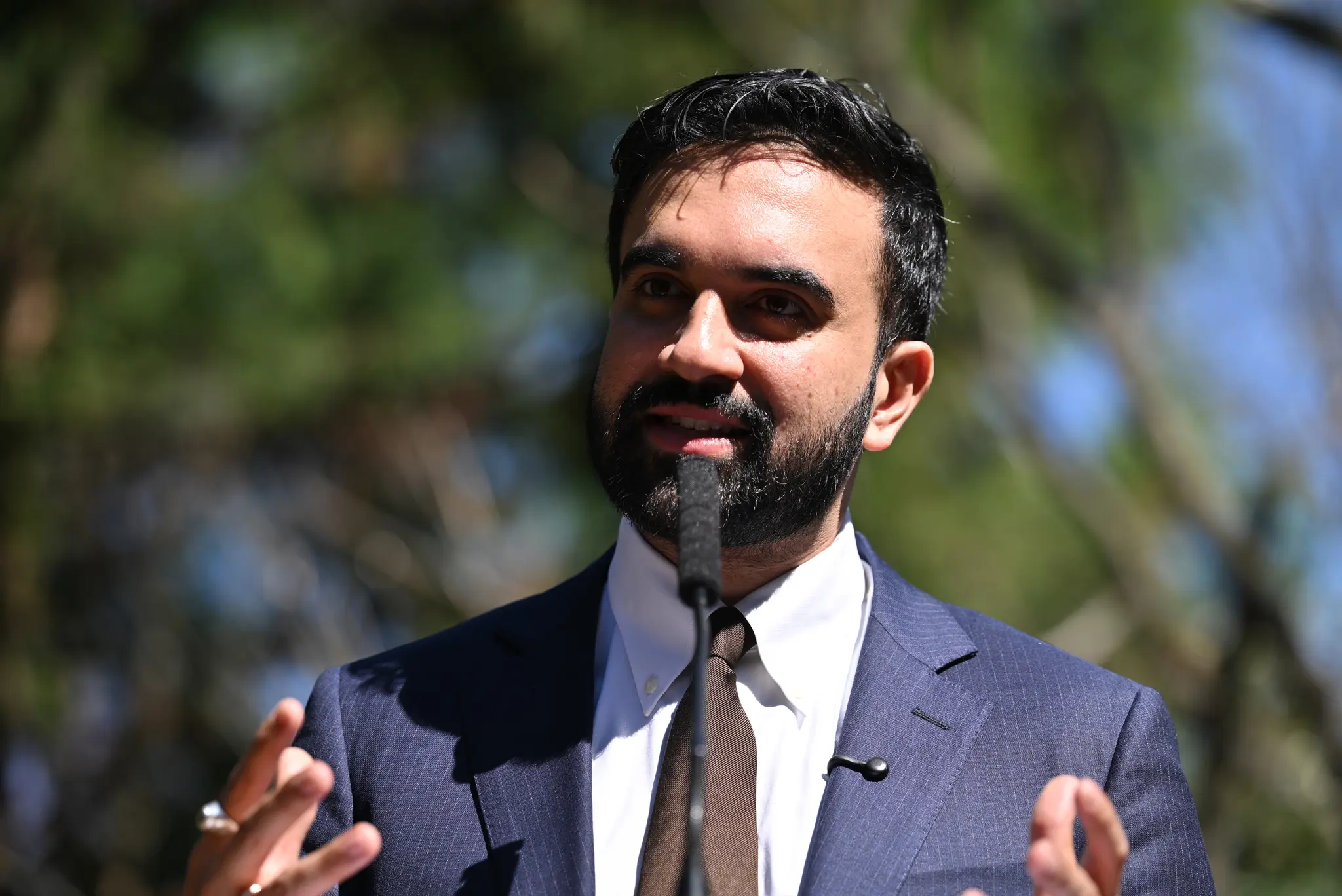 Mayor Zohran Mamdani speaks outdoors at a public event, wearing a suit and addressing the audience through a microphone.
