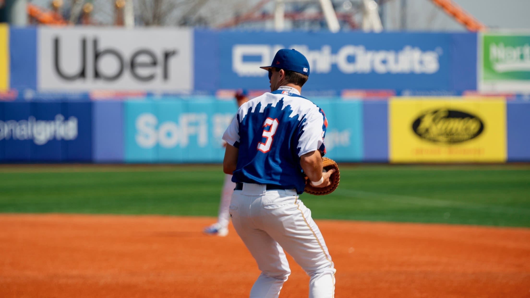 Brooklyn Cyclones infielder Trace Willhoite (#3) fields a ball on the infield with colorful outfield advertisements behind him.