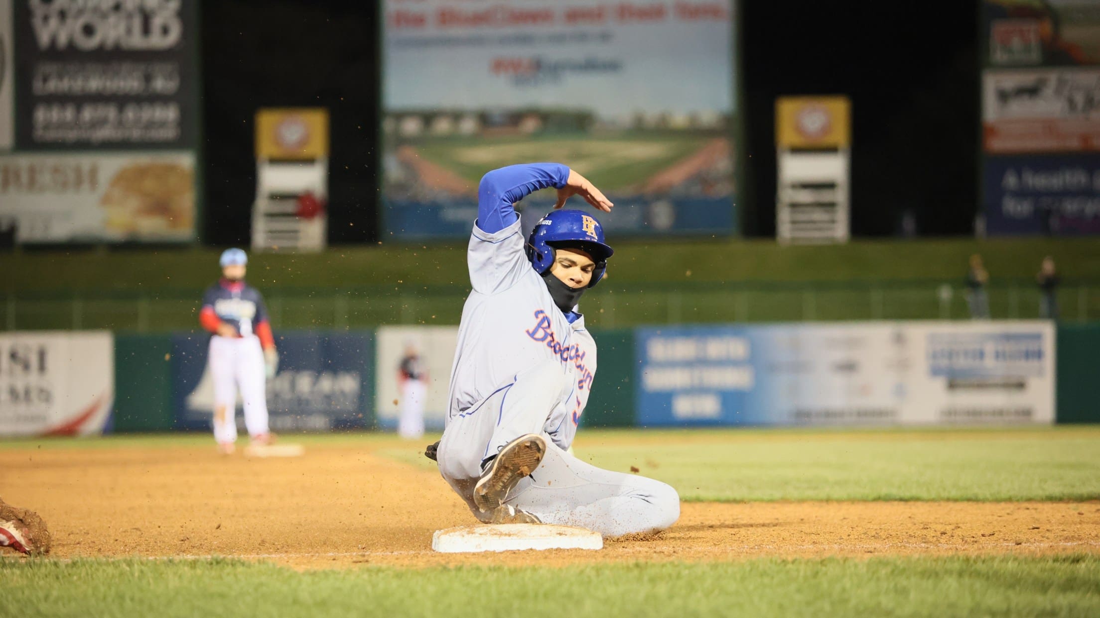 A Jersey Shore BlueClaws player slides into a base during a night game, kicking up dirt as a fielder looks on.