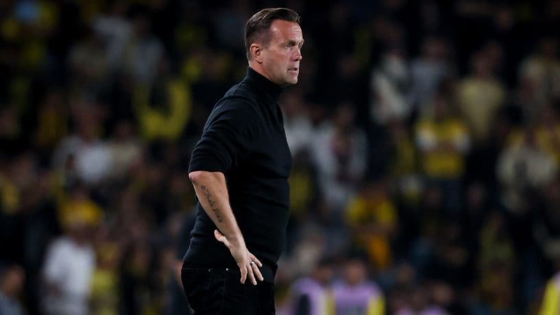 Ronny Deila stands on the sideline during a match, wearing a black turtleneck as he watches play unfold.