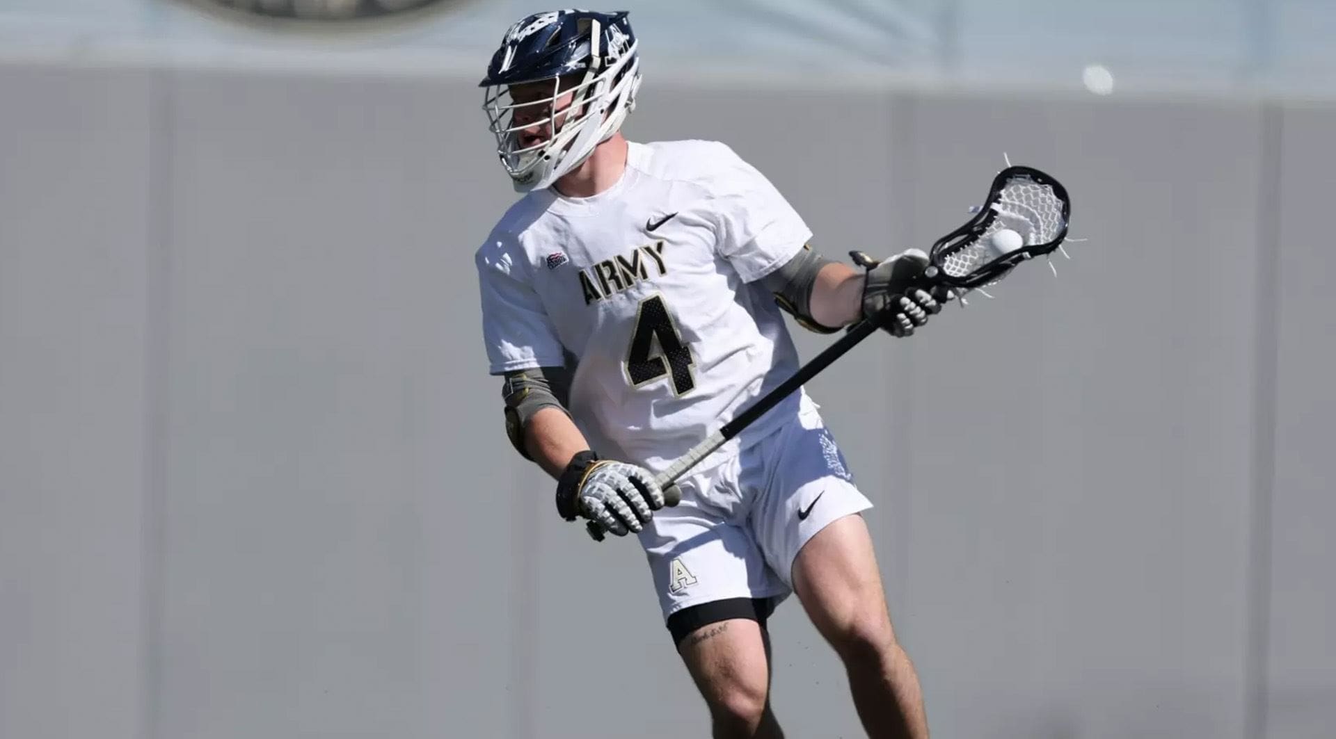 Army West Point midfielder Evan Plunkett carries the ball upfield during a game, wearing a white Army uniform and helmet.